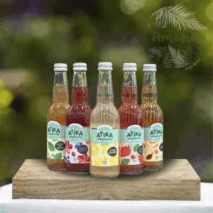 Five glass bottles of Atika kombucha arranged in a row on a wooden platform outdoors, each with a different colorful label flavor. Option: informative description for an article about beverages or a product lineup.