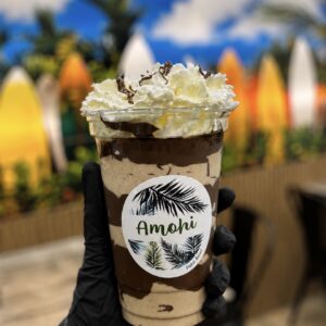 Screenshot - AMOHI Chocolate milkshake topped with whipped cream and chocolate drizzle in a clear cup labeled Amoohi, held by a gloved hand with a tropical backdrop.