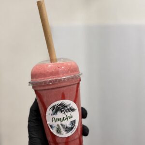 Screenshot - AMOHI Pink smoothie in a clear plastic cup with a dome lid and tan straw, labeled Amohi, held by a gloved hand against a light background.